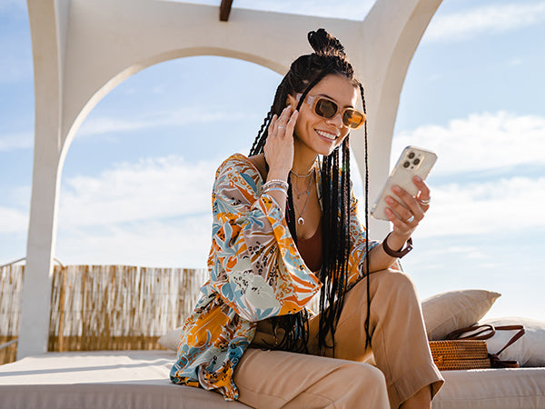 Stylish woman with braided hair and sunglasses holds white iPhone under beach arches, wearing orange patterned blouse and jewelry, lounging on cushions—2025 summer vibe with Ellis Edge phone case protection.