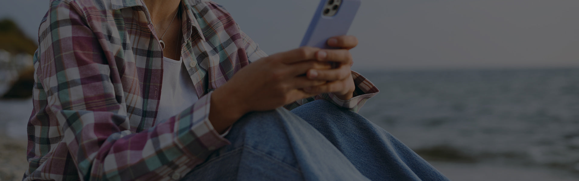 Woman in plaid shirt holding white iPhone on beach overlooking sea, relaxed pose—Tech Accessory Insights for Ellis Edge's 2025 phone cases & chargers.